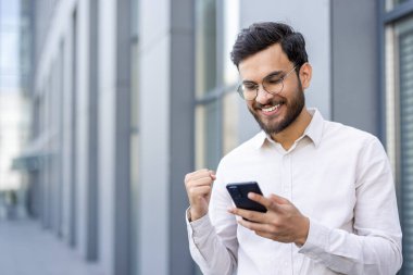 Gözlüklü neşeli bir adam telefonunda güzel haberler kutluyor. Modern bir binanın yakınında açık havada gülümsüyor..
