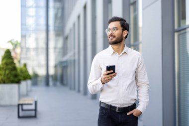 İş kıyafeti giymiş bir adam elinde akıllı bir telefonla modern bir ofis binasına doğru bakıyor..