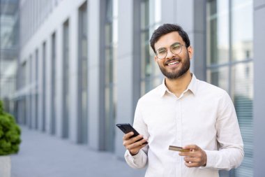 Gülümseyen bir adam modern bir binanın dışında telefon ve kredi kartı tutuyor, muhtemelen bir şeyler satın alıyor..