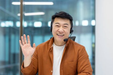 Neşeli Asyalı bir adam kameraya kulaklık takıyor, sıcak bir şekilde gülümsüyor, parlak bir ofis ortamında.