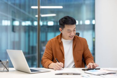 Asyalı bir adam masasında çalışıyor, hesap makinesi kullanarak gülümsüyor ve ofisteki bir belgeye yazı yazıyor..