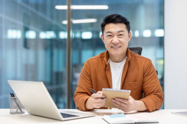 Masada oturan gülümseyen Asyalı bir adam elinde bir not defteri tutuyor ve modern bir ofiste çalışıyor..