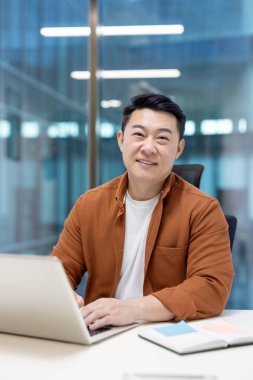 Modern ofis ortamında dizüstü bilgisayarla çalışırken gülümseyen Asyalı bir adam..