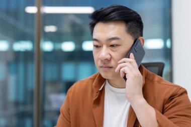 Kahverengi ceketli Asyalı bir adam, muhtemelen bir ofis ortamında, bir telefon görüşmesi yapıyor..