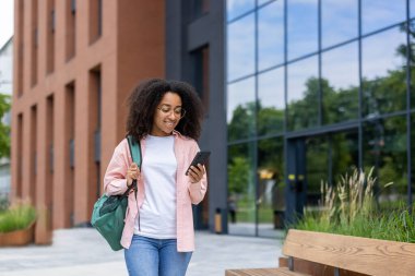 Bir Afrikalı Amerikalı öğrenci modern bir binanın yanında yürürken telefonuna bakarken gülümsüyor..