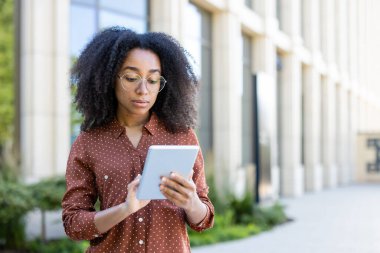Odaklanmış bir kadın gözlük takar, elinde tablet taşır ve arka planı modern bir binadır..
