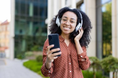 Yüzünde gözlük ve kulaklık olan gülümseyen Afro-Amerikalı bir kadın elinde akıllı bir telefonla bina arkası müziğinin keyfini çıkarıyor..