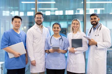 Doktorlar ve hemşireler de dahil olmak üzere çeşitli tıp uzmanları modern bir sağlık merkezinin içindeki kameraya gülümsüyor..