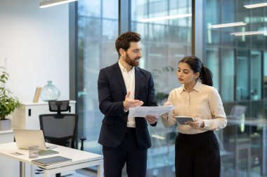 İki meslektaş, bir erkek ve bir kadın, modern ofis ortamında işbirliği yapıyorlar, belgeleri gözden geçiriyorlar ve bir tablet kullanıyorlar..