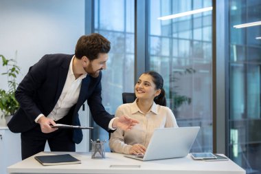 Bir işadamı, modern ofis ortamında dizüstü bilgisayar üzerinde çalışırken bir iş kadınına rehberlik eder ve tavsiyelerde bulunur..