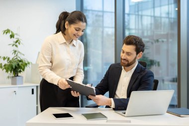 Bir kadın, modern ofis ortamında gülümseyen bir erkeğe bir belge sunar..