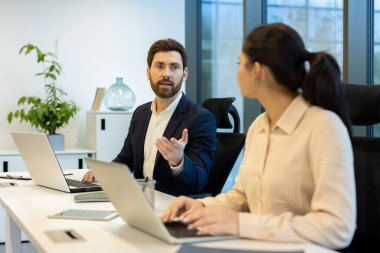 Modern ofis ortamında bir iş toplantısı başlar. Bir erkek dizüstü bilgisayarda çalışan bir kadınla konuşurken el kol hareketi yapar..