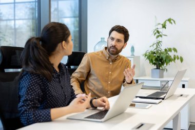 İki meslektaş, bir erkek ve bir kadın, modern ofis ortamında bir masada dizüstü bilgisayarlar üzerinde çalışıyorlar..