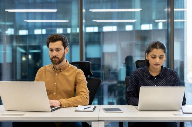 İki farklı meslektaş, modern bir ofiste dizüstü bilgisayarlarıyla çalışırlar ve görevlerine odaklanırlar..