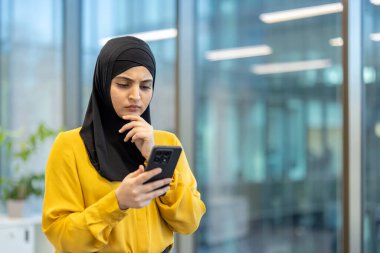Siyah tesettürlü ve sarı gömlekli bir kadın modern ofis ortamında telefonuna düşünceli bir şekilde bakar. Çenesinde bir eli var..