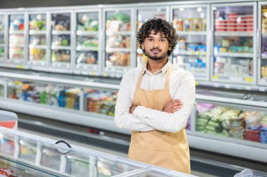 Önlük takan bir süpermarket çalışanı soğutuculu bölümün önünde kollarını kavuşturmuş duruyor..