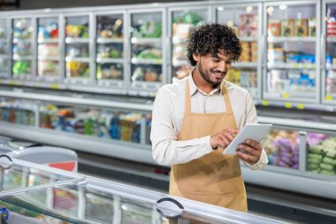 Önlük takan bir mağaza çalışanı, soğutuculu vitrinlerin önünde envanteri yönetmek için tablet kullanır ve çalışırken gülümser..
