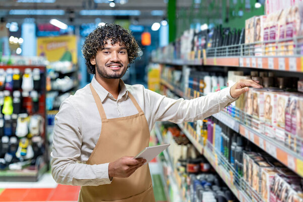 A smiling worker in a supermarket stands by shelves, checking inventory with a tablet while wearing an apron.