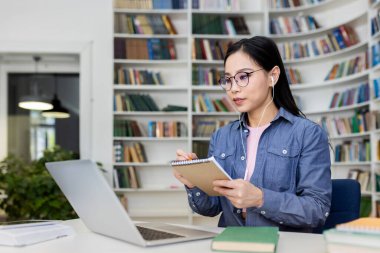 Kütüphanedeki Asyalı bir kadın video görüşmesi sırasında not alıyor. Yanında dizüstü bilgisayarı ve kitapları var..