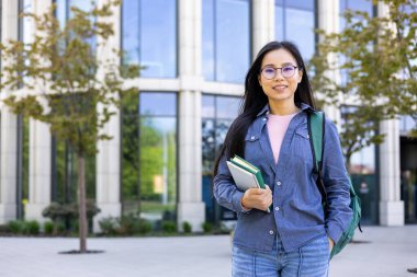 Kitapları ve sırt çantaları olan Asyalı bir öğrenci üniversite binasının önünde durup gülümsüyor..