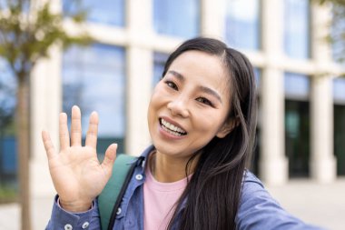 Sırt çantasıyla binanın önünde gülümseyen Asyalı kadın selamlarını ve pozitifliklerini bildiriyor..
