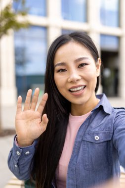 Asyalı bir kadın, kameraya sıcak bir gülümsemeyle dışarıda selfie çekerken el sallıyor..