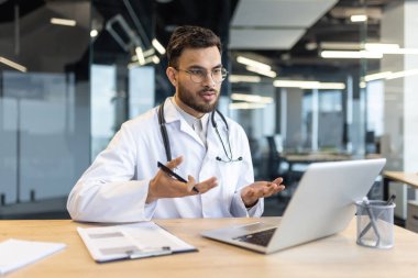 Beyaz önlüklü bir doktor, modern ofis ortamında video görüşmesi, danışmanlık veya telemedecine için dizüstü bilgisayar kullanır..