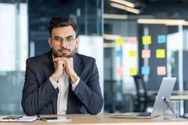 Gözlüklü, kendine güvenen bir işadamı modern ofis ortamında masasında poz veriyor..