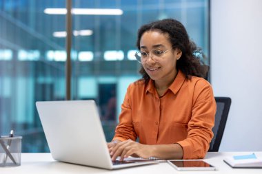 Genç bir iş kadını laptopuyla modern bir ofiste çalışıyor, gülümsüyor ve odaklanıyor. Sahne profesyonel ve üretken bir ortam gösteriyor..