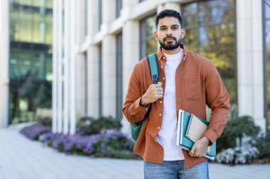 Bir öğrenci dışarıda duruyor, modern bir binanın önünde kitap ve sırt çantası tutuyor. Ders için hazır..