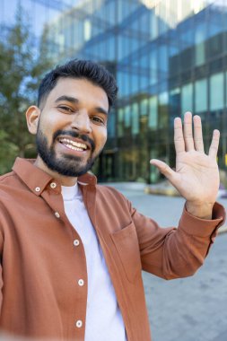 Modern bir binanın önünde kameraya el sallayan gülümseyen bir adam.