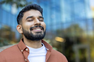 Gözleri kapalı gülümseyen bir Hintlinin yakın planda çekilmiş görüntüsü bulanık bir arkaplanda, açık havada eğleniyormuş gibi görünüyor..