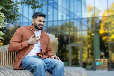 Göğüs ağrısı çeken, dışarıda oturan, muhtemelen kalp krizi ya da sağlık sorunu yaşayan bir adam..