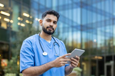 Önlüklü bir sağlık uzmanı modern bir binanın önünde açık havada tablet kullanıyor. Steteskop takıyor..