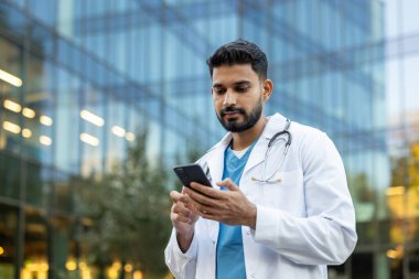 Beyaz önlüklü bir doktor modern bir binanın dış yüzeyine karşı telefon kullanıyor. Cihazına odaklanmış görünüyor, muhtemelen iletişim kuruyor..