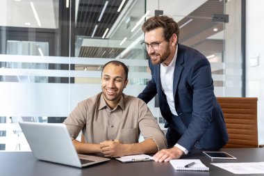 İki meslektaş bir projede işbirliği yapıyor, biri bir dizüstü bilgisayarla masada otururken, diğeri destek vermek için öne eğiliyor.