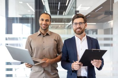 İki işadamı bir ofis ortamında güven içinde poz veriyor, biri dizüstü bilgisayar tutuyor, diğeri de bir dosya, ikisi de gülümsüyor..