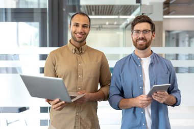 Modern bir ofis ortamında elinde bir dizüstü bilgisayar ve tablet olan iki farklı meslektaş gülümsüyor. Arkadaş canlısı ve profesyonel görünüyorlar..