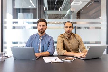Masada dizüstü bilgisayarlar üzerinde çalışan iki işadamı modern ofis ortamında kameraya bakıyor..