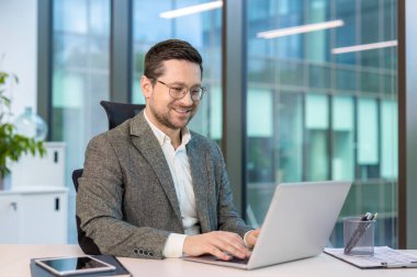Gülümseyen genç işadamı ofiste bir masada takım elbiseyle oturuyor ve bir dizüstü bilgisayar üzerinde çalışıyor..