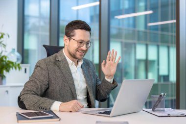 Gülümseyen Kafkasyalı genç ofis çalışanı masasında oturmuş, internette konuşuyor ve dizüstü bilgisayar kamerasını selamlıyor..
