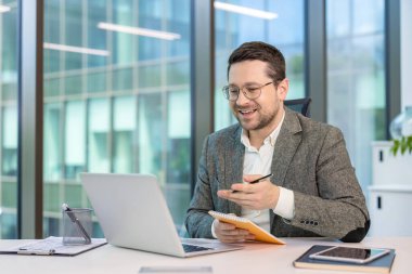 Ofisindeki iş yerinde oturmuş bilgisayarla konuşan takım elbiseli gülümseyen genç bir işadamı. Not defterine notlar alıyor..