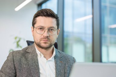 İş yerindeki ofisinde dizüstü bilgisayarla oturan ve kameraya ciddi ciddi bakan genç bir erkek işadamının yakın plan portresi..