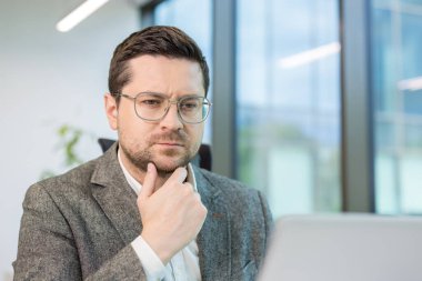 Ofiste oturan ciddi bir gencin yakın plan fotoğrafı, özenle dizüstü bilgisayara bakıyor, bir soruyu ve problemi çözüyor..