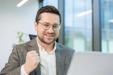 Ofiste oturan, başarıyla sevinen, zafer jesti yapan ve dizüstü bilgisayar ekranına bakan genç bir adamın yakın çekimi..