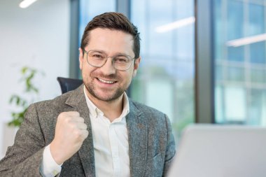 Genç ve başarılı bir iş adamının portresi ofiste dizüstü bilgisayarın önünde oturuyor, seviniyor, zafer jestini eliyle gösteriyor ve kameraya gülümsüyor..