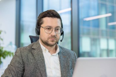 Takım elbiseli ve kulaklıklı genç bir adamın ofiste dizüstü bilgisayarla çalışırken çekilmiş yakın plan fotoğrafı..