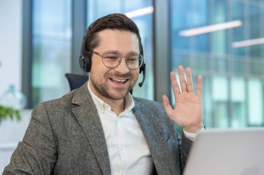 Bilgisayarın başındaki ofiste çalışan ve kameraya el sallayan, kulaklık takmış gülümseyen genç bir adamın yakın plan fotoğrafı..