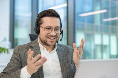Genç bir erkek ofis çalışanı ofiste bir masada oturuyor, kulaklık takıyor ve dizüstü bilgisayarla konuşuyor, elleriyle işaret ediyor..