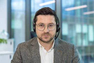 İş yerinde kulaklıkla oturan ve kameraya dikkatlice bakan ciddi bir erkek ofis çalışanı ve asistanının yakın plan portresi..